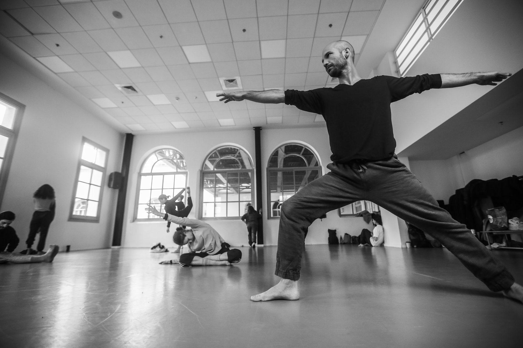 Tal in a wide stance with arms open, students moving behind him in the studio